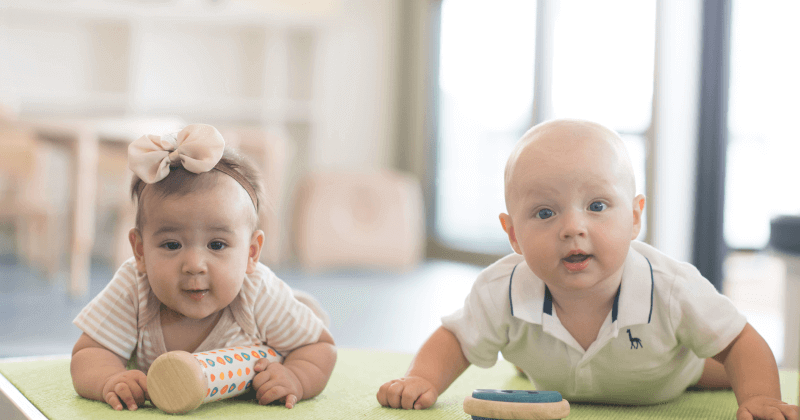 A smiling infant lying on their tummy on a soft mat in a bright, modern infant care environment, engaging in tummy-time play. - Australian International School (AIS)