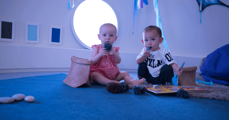 Baby sitting on a blue cushioned floor, reaching out to hold a soft toy, in a calm, sensory-rich learning space.- Australian International School (AIS)